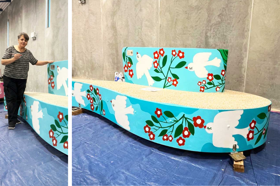 Woman standing next to bench she painted with white doves next to red flowers on a bright sky