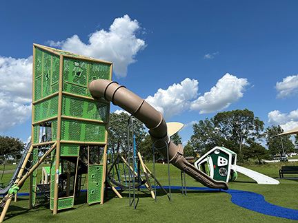 Tall green climbing structure with slide coming down to turf and camper inspired play equipment