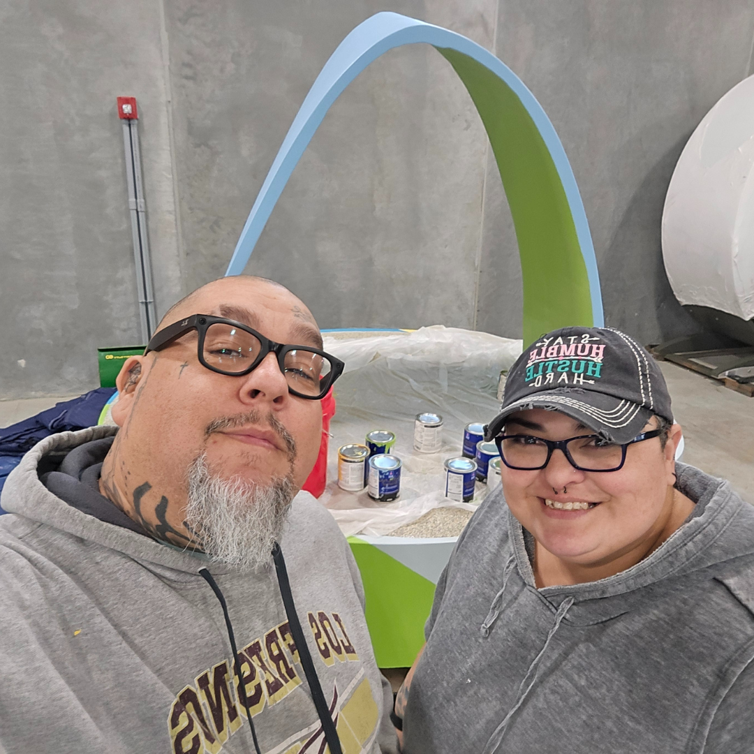 Selfie of man with glasses and short beard and woman with short hair in front of bench they painted